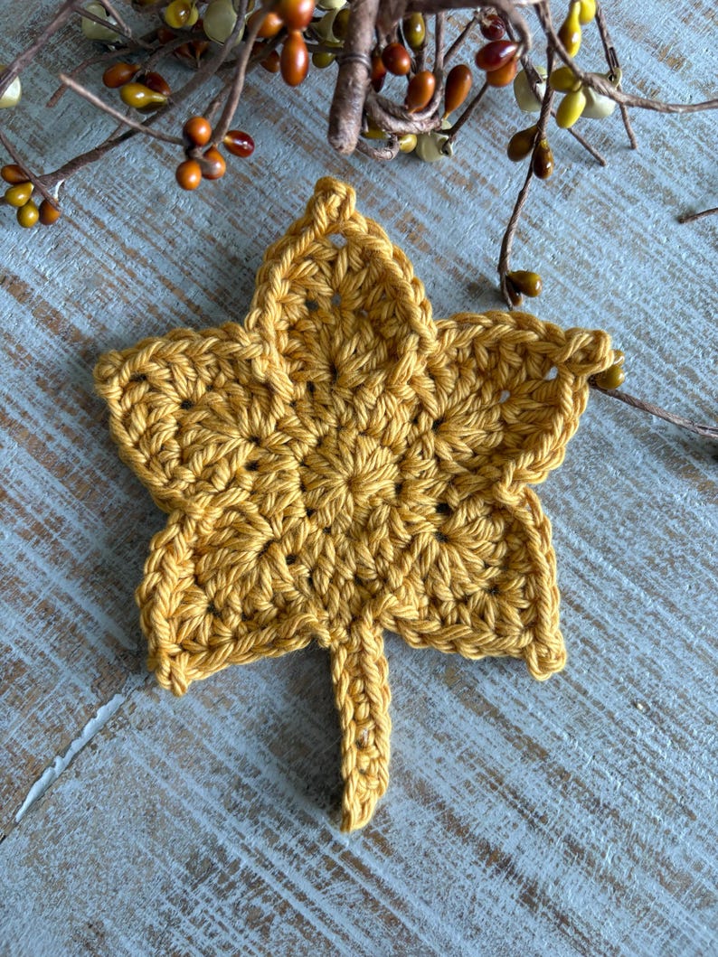 May include: A handmade, golden yellow crochet leaf. The leaf is detailed with visible stitches and a textured surface. The leaf is placed on a light blue wooden surface, with fall-themed decorative branches in the background.