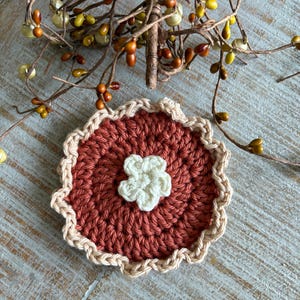 May include: A handmade crochet coaster with a pie-like design. The coaster features a rust-colored center with a cream-colored flower in the middle. The edge is trimmed with a cream-colored scalloped border. Decorative fall-themed branches with berries and bells are in the background.