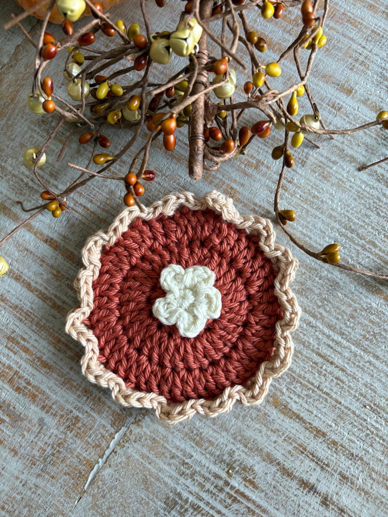 May include: A handmade crochet coaster with a pie-like design. The coaster features a rust-colored center with a cream-colored flower in the middle. The edge is trimmed with a cream-colored scalloped border. Decorative fall-themed branches with berries and bells are in the background.