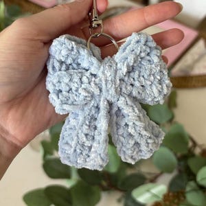 May include: A light blue, fluffy bow-shaped keychain. The bow is made of a soft, textured material and is attached to a silver-colored metal clasp and keyring. The keychain is held up against a blurred background of greenery and a pink tray.