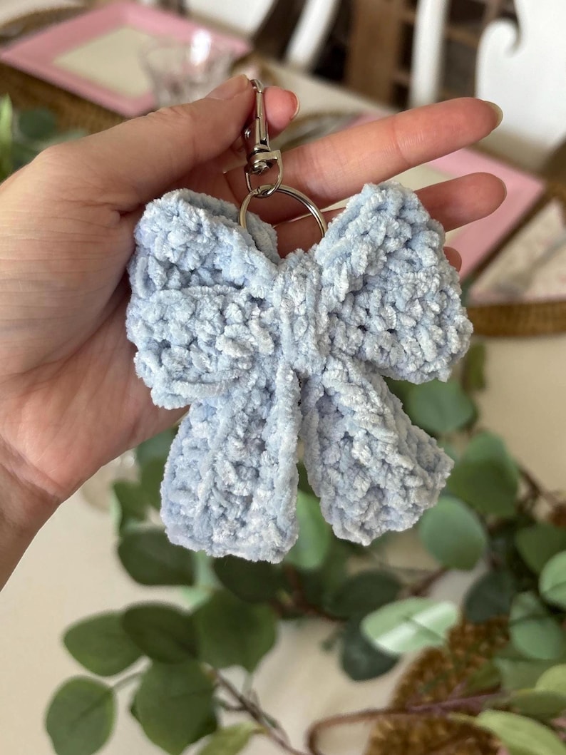 May include: A light blue, fluffy bow-shaped keychain. The bow is made of a soft, textured material and is attached to a silver-colored metal clasp and keyring. The keychain is held up against a blurred background of greenery and a pink tray.