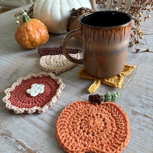 May include: A brown ceramic mug sits on a yellow leaf-shaped crochet coaster. Other crochet coasters include a pumpkin, a pie, and a leaf. Decorative pumpkins and gourds are in the background, creating a fall theme.