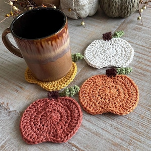 Pode incluir: Uma caneca de cerâmica marrom repousa sobre um descanso de copo de abóbora amarelo de crochê. São exibidos três descansos de copo de abóbora de crochê adicionais em branco, laranja e vermelho-alaranjado. Cada descanso de copo tem uma haste marrom e detalhes de folhas verdes.