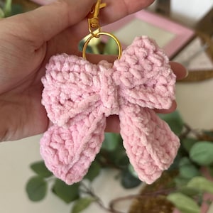 May include: A light pink, crocheted bow keychain with a gold-colored clasp and ring. The bow is made of a soft, textured yarn. The keychain is a decorative accessory.