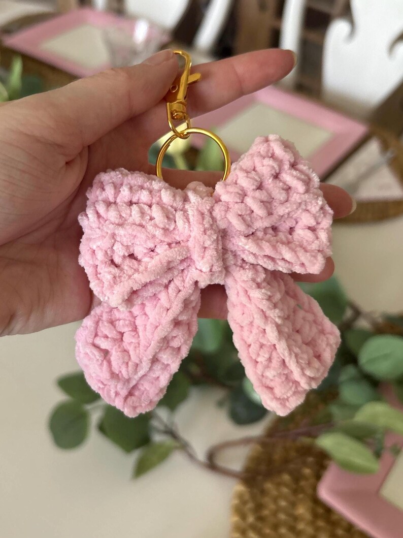 May include: A light pink, crocheted bow keychain with a gold-colored clasp and ring. The bow is made of a soft, textured yarn. The keychain is a decorative accessory.