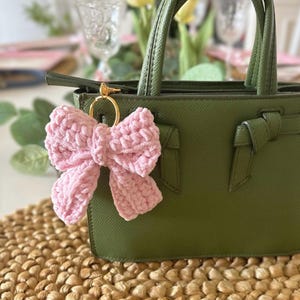 May include: A light pink, plush bow keychain attached to a green handbag with gold hardware. The bow is made of a soft, textured material. The handbag has a structured design with top handles.
