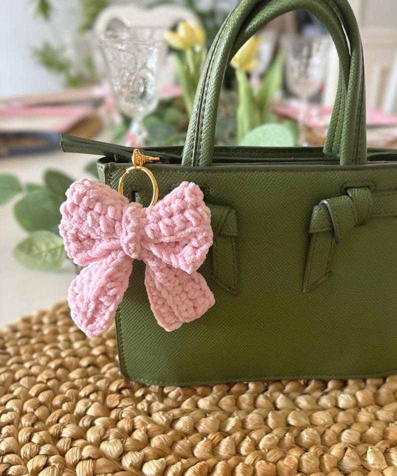 May include: A light pink, plush bow keychain attached to a green handbag with gold hardware. The bow is made of a soft, textured material. The handbag has a structured design with top handles.