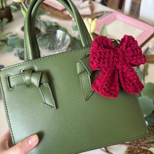 May include: A green leather handbag with a top handle and a decorative bow detail on the front. The bag has a rectangular shape and a textured surface. A red crocheted bow keychain is attached to the bag.