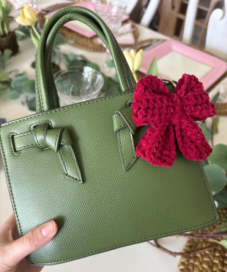 May include: A green leather handbag with a top handle and a decorative bow detail on the front. The bag has a rectangular shape and a textured surface. A red crocheted bow keychain is attached to the bag.