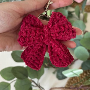May include: A handmade, crimson red crochet bow keychain. The bow is fluffy and textured, with a silver-colored metal clasp. The keychain is held up against a background of green foliage and a blurred pink frame.