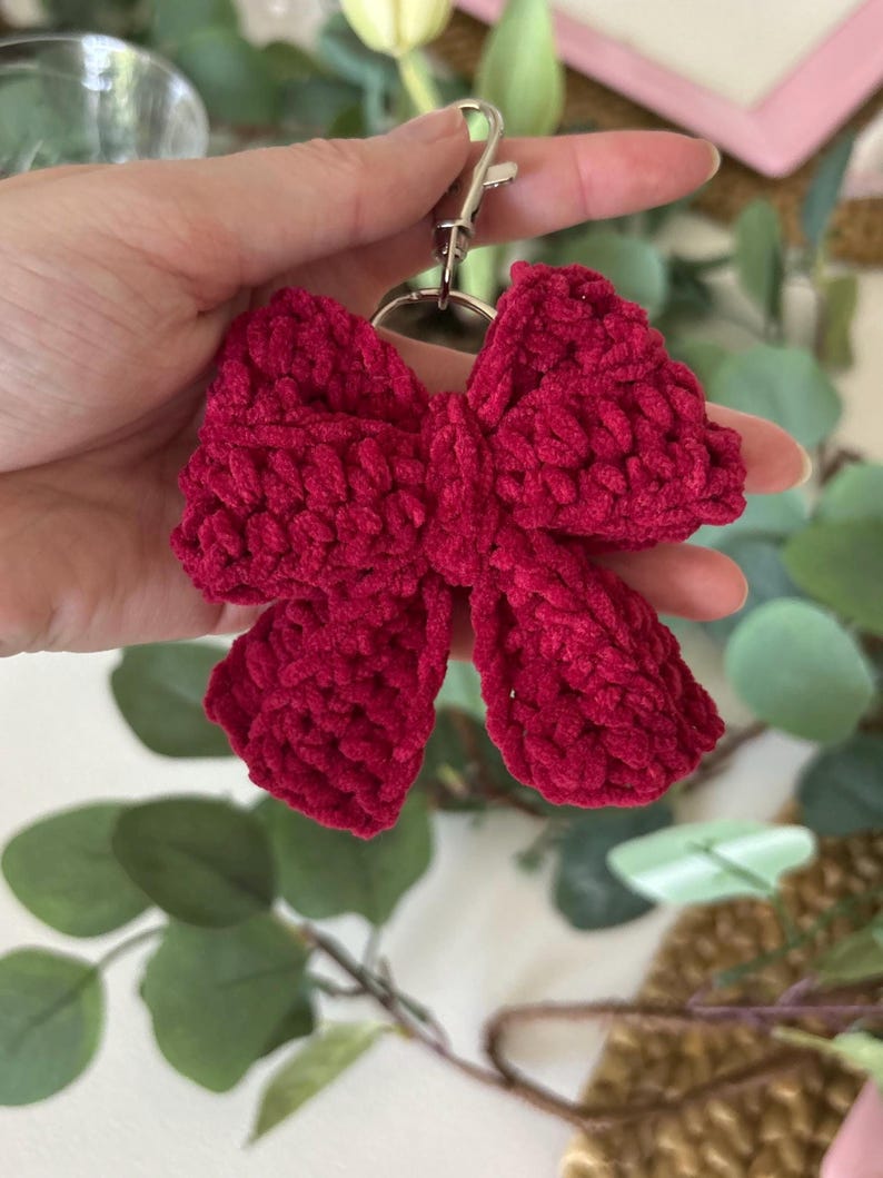 May include: A handmade, crimson red crochet bow keychain. The bow is fluffy and textured, with a silver-colored metal clasp. The keychain is held up against a background of green foliage and a blurred pink frame.