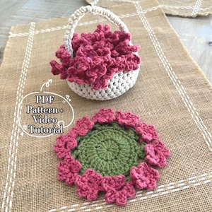 Op de afbeelding: Een gehaakte mand gevuld met roze bloemen en een bijpassende onderzetter. De mand is wit met een beige handvat. De onderzetter heeft een groen midden en roze bloemenrand. De afbeelding bevat de tekst "PDF Pattern + Video Tutorial."