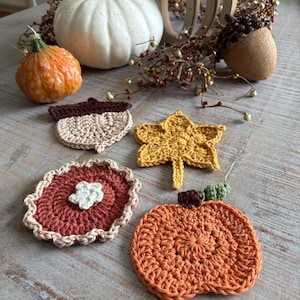 May include: A collection of handmade crochet fall-themed items. Includes an orange pumpkin, a yellow leaf, a brown acorn, and a pie-shaped coaster. Decorative pumpkins and fall foliage are in the background. The items are arranged on a wooden surface.