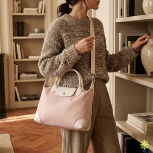 Puede incluir: Un bolso rosa claro con detalles en cuero blanco, un asa superior y una correa para el hombro. El bolso es rectangular con un cierre de solapa. La persona lleva un su&eacute;ter y pantalones marrones.