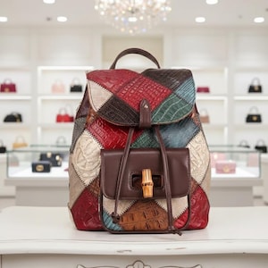 Può includere: Uno zaino in pelle patchwork multicolore con colori come rosso, marrone, verde e bianco. La borsa ha una chiusura a patta e una piccola tasca frontale con chiusura a forma di bambù. Lo zaino è appoggiato su una superficie bianca.