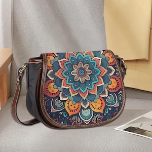 Puede incluir: Bolso tipo sillín negro con un colorido diseño de mandala en la solapa. El bolso presenta ribetes de cuero marrón, una correa de hombro marrón y herrajes dorados. El diseño del mandala incluye tonos de azul, naranja y turquesa.