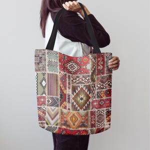 May include: A tote bag featuring a patchwork design with geometric patterns in red, brown, beige, and green. It has black handles and a rectangular shape, resembling a vintage rug. The bag is held by a person.