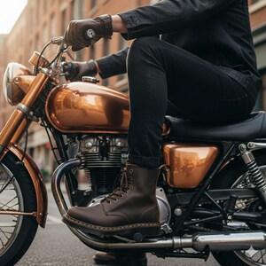 May include: A person is seated on a vintage copper-colored motorcycle, wearing black jeans and brown leather boots. The boots are laced up and have a classic design. The person is also wearing brown leather gloves.
