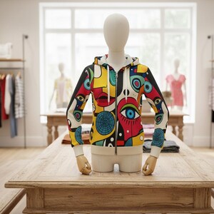 Op de afbeelding: Een zip-up hoodie met een levendig, abstract ontwerp met gezichten, ogen en geometrische vormen in rood, geel, blauw en zwart. De hoodie heeft een witte binnenkant en manchetten, tentoongesteld op een mannequin in een kledingwinkel.