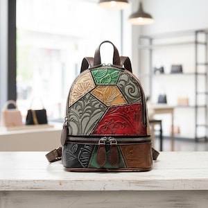 Puede incluir: Mochila de cuero multicolor con asa superior y correas ajustables. La bolsa presenta patrones florales y geométricos en relieve en tonos verdes, rojos, marrones y negros. La mochila tiene un compartimento principal con cremallera y un bolsillo más pequeño con cremallera.