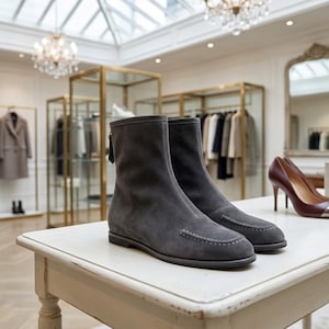 Könnte beinhalten: Ein Paar graue Wildleder-Ankle-Boots mit Reißverschlussdetail am Rücken und weißen Nähten an der Spitze. Die Stiefel werden auf einem weißen Tisch in einer Boutique präsentiert, mit bordeauxfarbenen High Heels im Hintergrund.