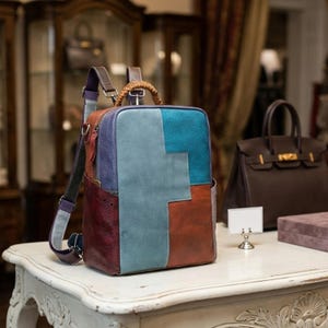 Puede incluir: Una mochila de cuero multicolor con un diseño de patchwork. La mochila presenta una combinación de paneles azules, morados, grises y marrones. Tiene correas ajustables, un asa superior y un bolsillo frontal. La bolsa está sobre una mesa blanca.