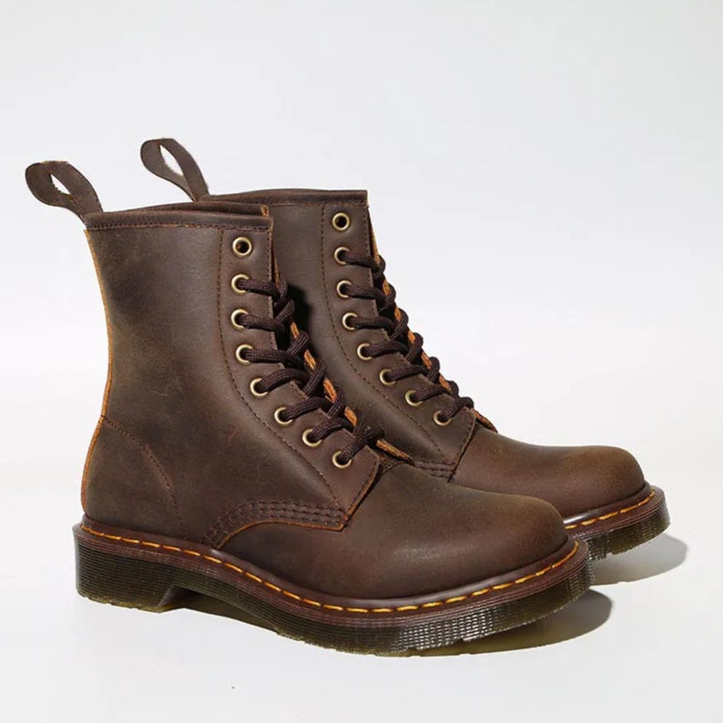 May include: A pair of brown leather boots with a lace-up design. The boots have a rugged, worn appearance, with visible stitching and metal eyelets. The soles are dark brown with yellow stitching. The boots have pull tabs at the top.