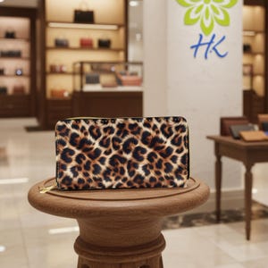 Puede incluir: Una cartera con estampado de leopardo y cremallera dorada se exhibe sobre una pequeña mesa de madera. La cartera presenta un patrón de manchas marrones y negras sobre un fondo beige. El fondo muestra una tienda con otros bolsos.
