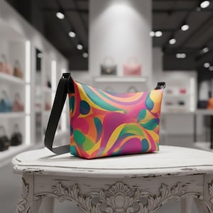 May include: A colorful crossbody bag with a vibrant, abstract pattern in shades of pink, yellow, green, and blue. The bag has a black strap and is displayed on a white, ornate table. The background shows a store with other bags.