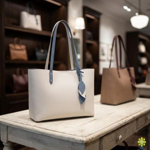 May include: A cream-colored tote bag with light blue handles and a matching leaf-shaped charm. The bag is displayed on a distressed white wooden table, with other bags visible in the background on shelves.