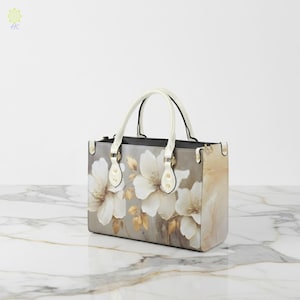 May include: A rectangular handbag with a floral design. The bag features white flowers and gold leaves against a grey background. It has white handles and gold-coloured hardware. The bag is approximately 30 cm wide.