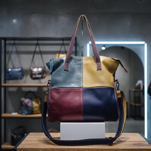 Peut inclure: Sac fourre-tout en cuir multicolore avec un motif patchwork. Il présente des panneaux bleus, jaunes, bordeaux et marine. Il possède une bandoulière et des poignées supérieures avec un accent violet. Le sac est présenté sur une surface en bois.