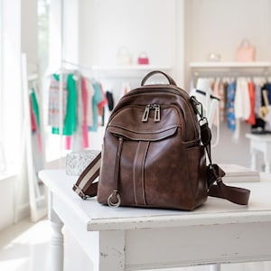 Può includere: Uno zaino in pelle marrone con maniglia superiore, tasca frontale e spallacci regolabili. Lo zaino ha cerniere argentate e un design cucito bianco. È appoggiato su un tavolo bianco in una boutique.