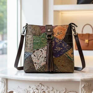 Könnte beinhalten: Eine mehrfarbige Patchwork-Leder-Umhängetasche mit geprägten Blumen- und geometrischen Mustern. Sie hat einen langen braunen Riemen und eine Quaste. Die Tasche steht auf einer weißen Marmoroberfläche. Die Tasche ist ca. 25 cm hoch.