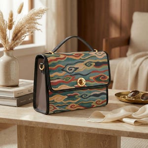 May include: A structured handbag with a colorful cloud pattern in shades of teal, red, and yellow. The bag has a black handle and trim, a gold-tone clasp, and a rectangular shape. The bag sits on a marble table.