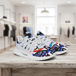 May include: White athletic shoes with colourful abstract designs. The shoes feature a white sole and laces, with a mix of blue, red, and black patterns on the upper. The shoes are displayed on a light-coloured wooden surface.