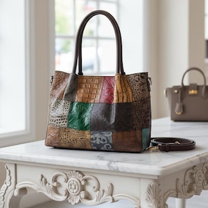 May include: A patchwork leather tote bag with a brown handle and a detachable shoulder strap. The bag features a variety of textures and colours, including brown, green, red, and tan. The bag is on a white marble table.