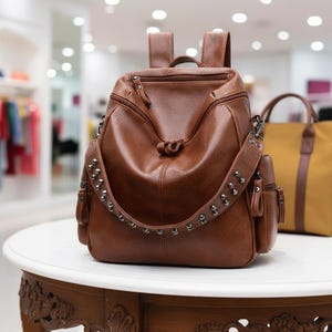 Op de afbeelding: Een bruine leren rugzak met een schouderband met studs en meerdere vakken. De tas heeft een handvat aan de bovenkant en verstelbare bandjes. Hij staat op een witte tafel met een decoratieve houten voet. Een andere tas is zichtbaar op de achtergrond.