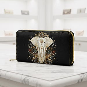 Pode incluir: Carteira preta com zíper, com um design detalhado de elefante branco, detalhes dourados e elementos florais. A carteira tem um zíper e costuras douradas.