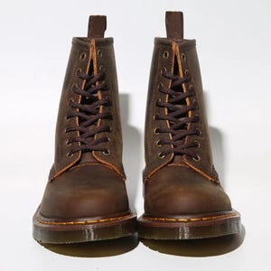 May include: A pair of brown leather boots with a lace-up design. The boots have a rugged, worn appearance, with visible stitching and a sturdy sole. The boots have a pull tab at the top and are set against a white background.