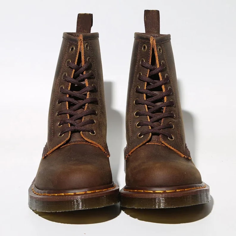 May include: A pair of brown leather boots with a lace-up design. The boots have a rugged, worn appearance, with visible stitching and a sturdy sole. The boots have a pull tab at the top and are set against a white background.