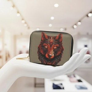 Puede incluir: Una pequeña cartera rectangular con cierre de cremallera. La cartera presenta una ilustración detallada de la cabeza de un lobo en tonos rojos y negros sobre un fondo gris. La cartera se sostiene en una mano blanca.