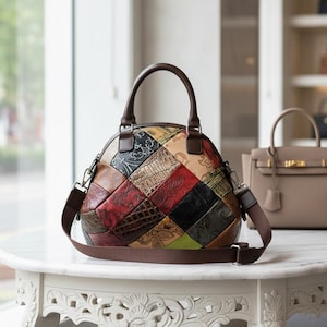 Może przedstawiać: Wielokolorowa skórzana torebka patchwork z brązową rączką i paskiem na ramię. Torba ma różne kolory i tekstury, w tym czerwony, zielony i czarny. Torba stoi na białym stole.