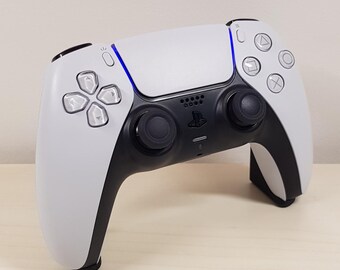Puede incluir: Un mando de videojuegos inalámbrico blanco y negro con un diseño simétrico. El mando cuenta con un panel táctil, botones direccionales y palancas analógicas. Una luz azul acentúa el borde superior. Diseñado para jugar.