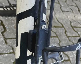 May include: A close-up of a black bicycle frame with white detailing and a black water bottle cage. The cage is attached to the frame, designed to hold a water bottle. The bike is on a grey surface.