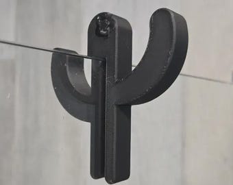 May include: A black metal cactus-shaped shower door hook. The hook is mounted on a glass shower door. The cactus design has two curved arms and a vertical stem. The hook is designed to hold a towel or other bathroom items.