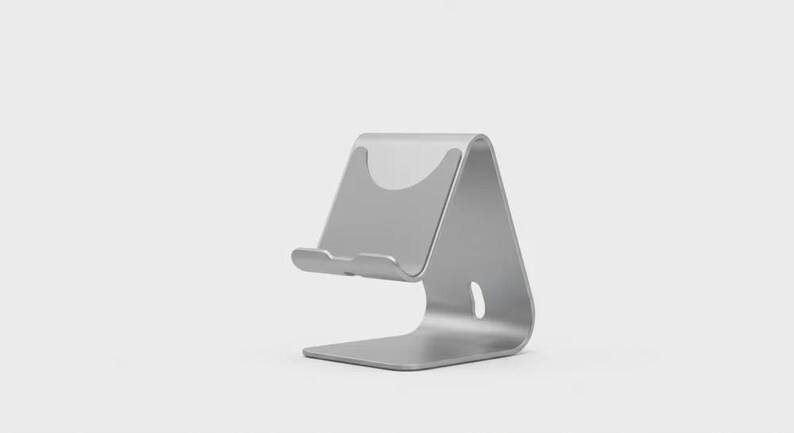 May include: A silver-colored phone stand with a curved design. The stand has a base, a back support, and a small ledge to hold a phone. The stand is made of metal and has a smooth finish.