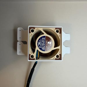 May include: A computer fan with a beige and brown fan blade design, mounted in a white frame. The fan has a black cable and the brand name "Noctua" is visible on the fan.