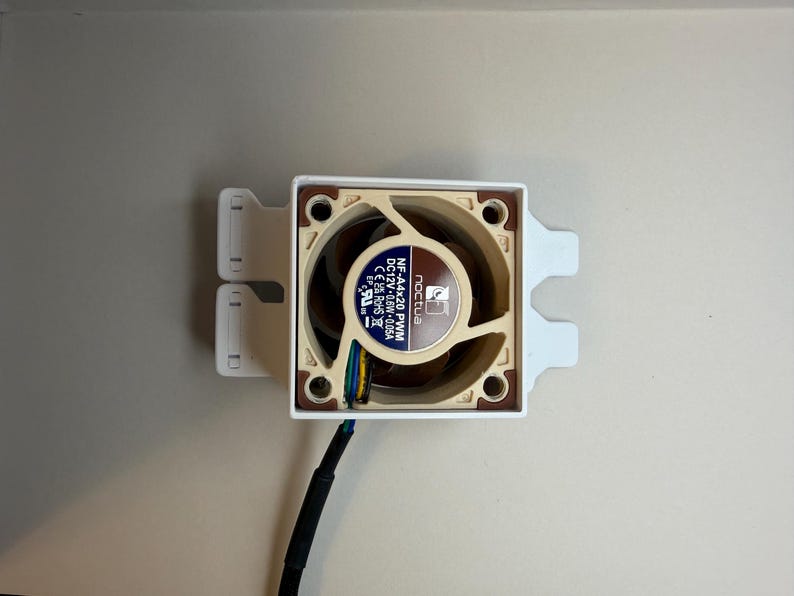 May include: A computer fan with a beige and brown fan blade design, mounted in a white frame. The fan has a black cable and the brand name "Noctua" is visible on the fan.