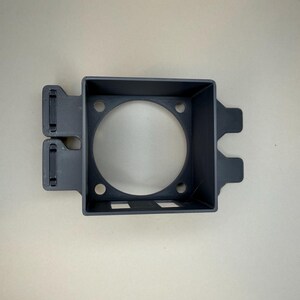 May include: A dark grey, rectangular plastic component with a circular aperture in the centre. It features two rectangular cutouts and several small holes. Two tabs extend from the side, against a light background.